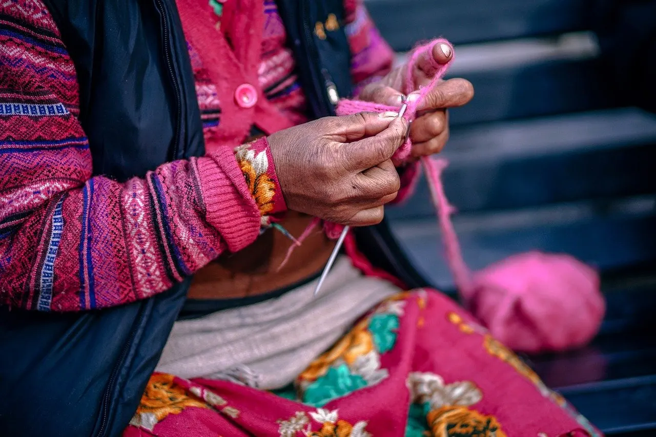 Faltige Hände einer älteren Frau stricken mit rosa Wolle, rechts ein Wollknäuel, darunter ein bunt gemusterter Rock