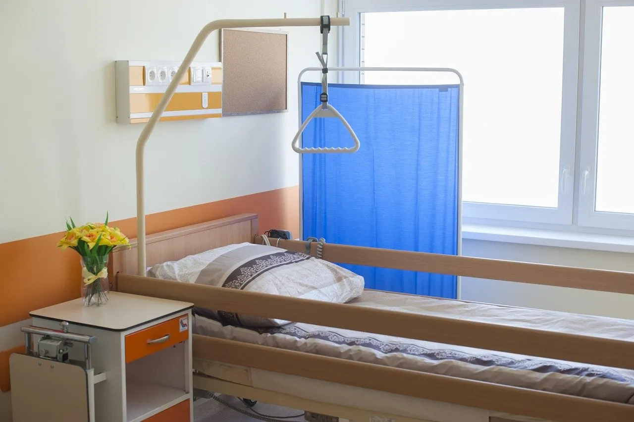 Leeres Pflegebett mit Seitengittern, Kopfkissen und Aufrichthilfe am Galgen, daneben ein Nachttisch mit gelben Blumen