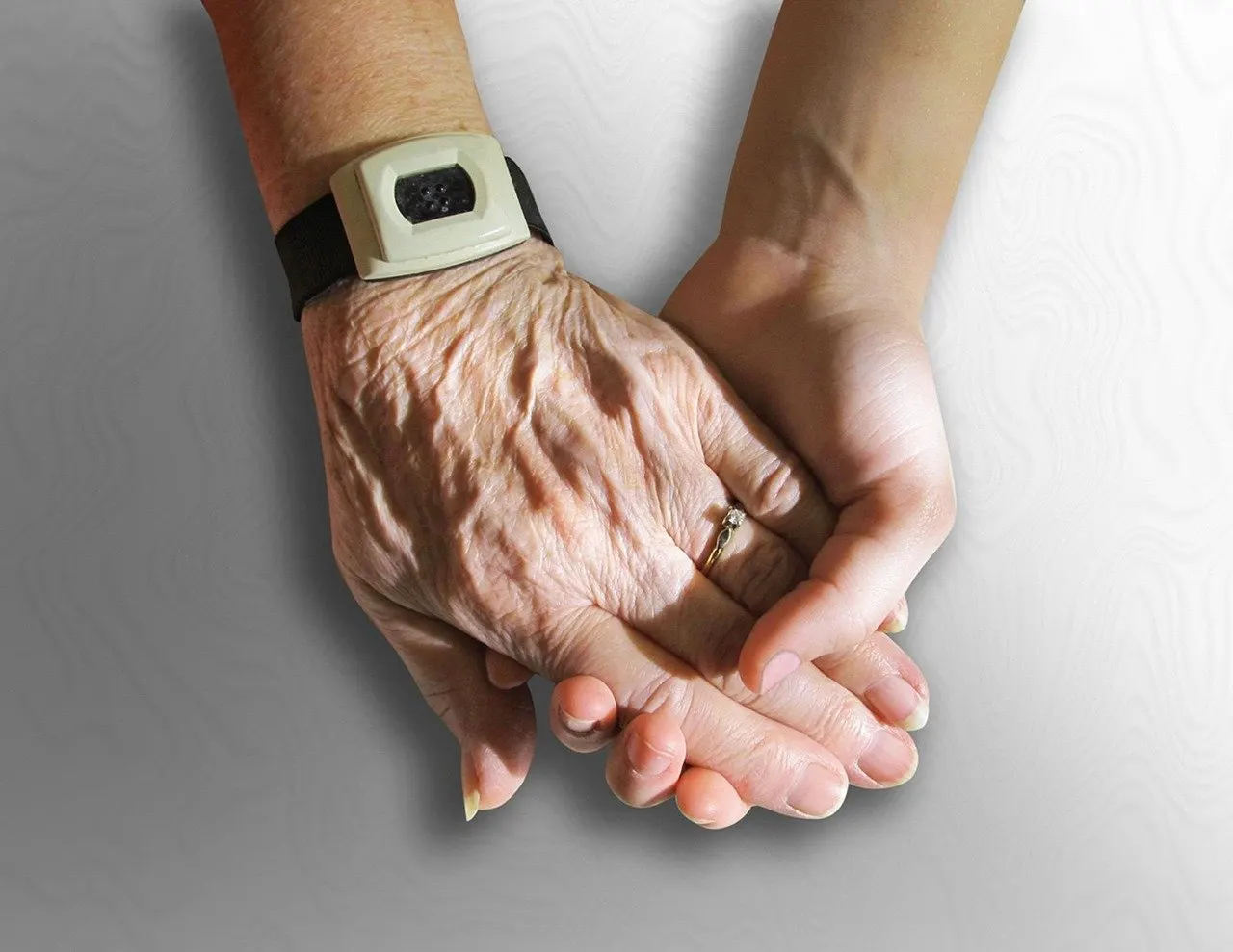 Eine faltige ältere Hand mit Ring und Hausnotruf-Armband liegt in einer jüngeren Hand, vor hellgrauem Hintergrund