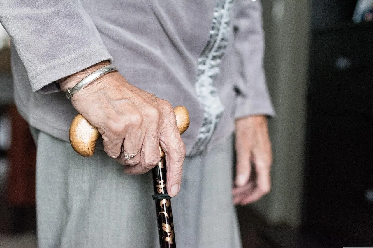 Hand einer älteren Frau mit Armreif und Ring auf dem Holzgriff eines Gehstocks, im Hintergrund ein Wohnraum