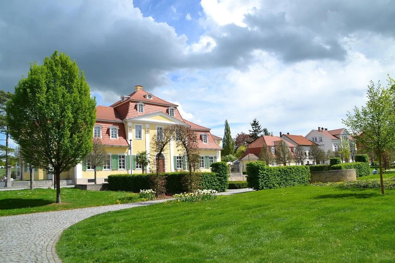 Gelbes Barockgebäude mit rotem Dach am Rand eines Kurparks mit Rasen, gepflastertem Weg und jungen Bäumen unter bewölktem Himmel