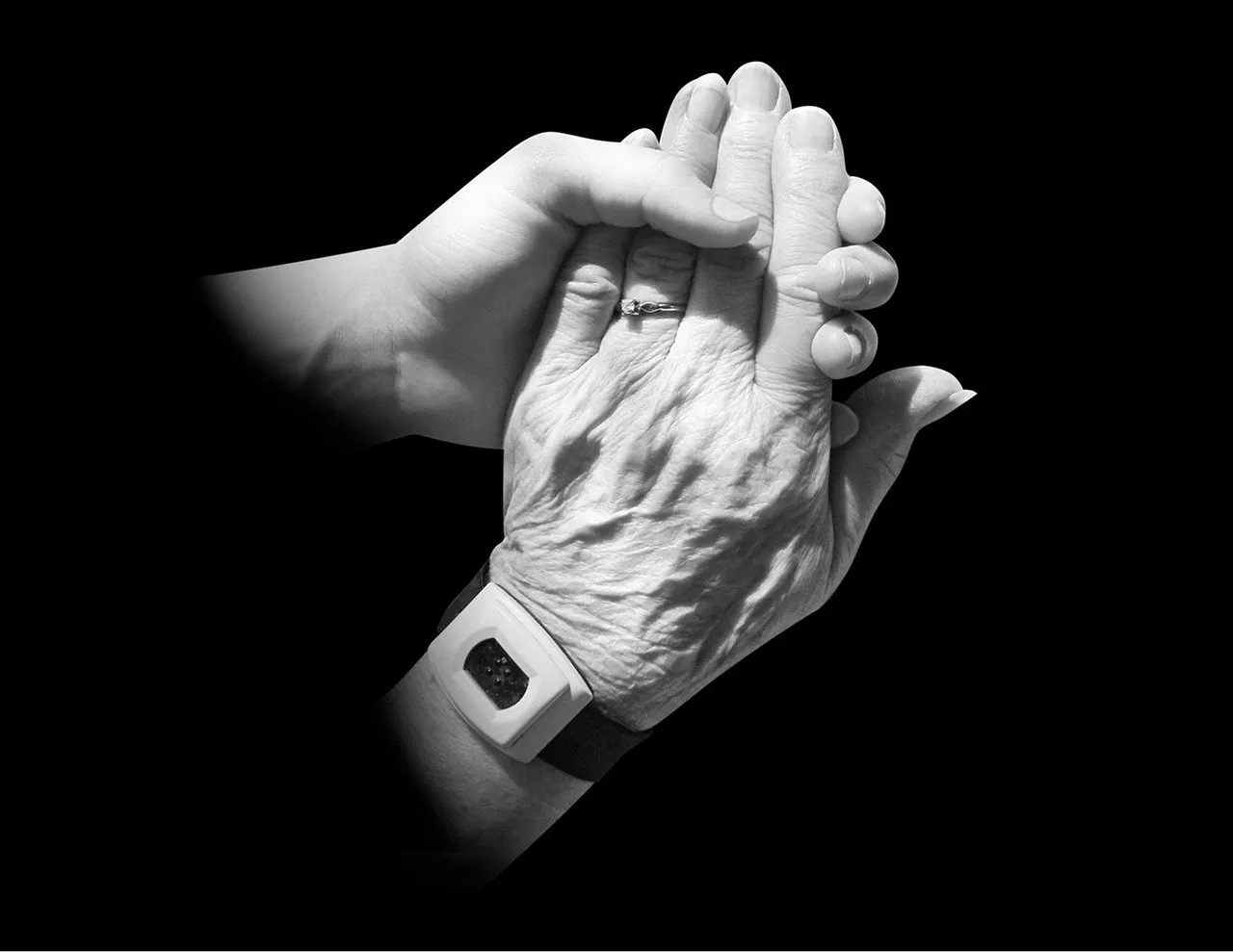 Schwarz-Weiß-Foto: Eine jüngere Hand umfasst eine faltige ältere Hand mit Ring, am Handgelenk ein helles Armband mit Knopf