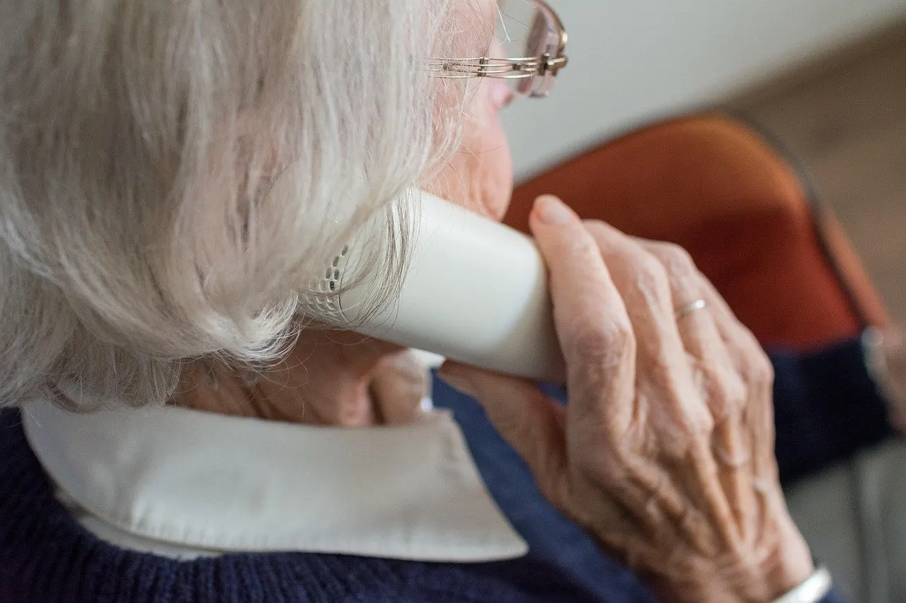 Ältere Frau mit grauem Haar und Brille, von hinten seitlich gesehen, hält einen weißen Telefonhörer ans Ohr