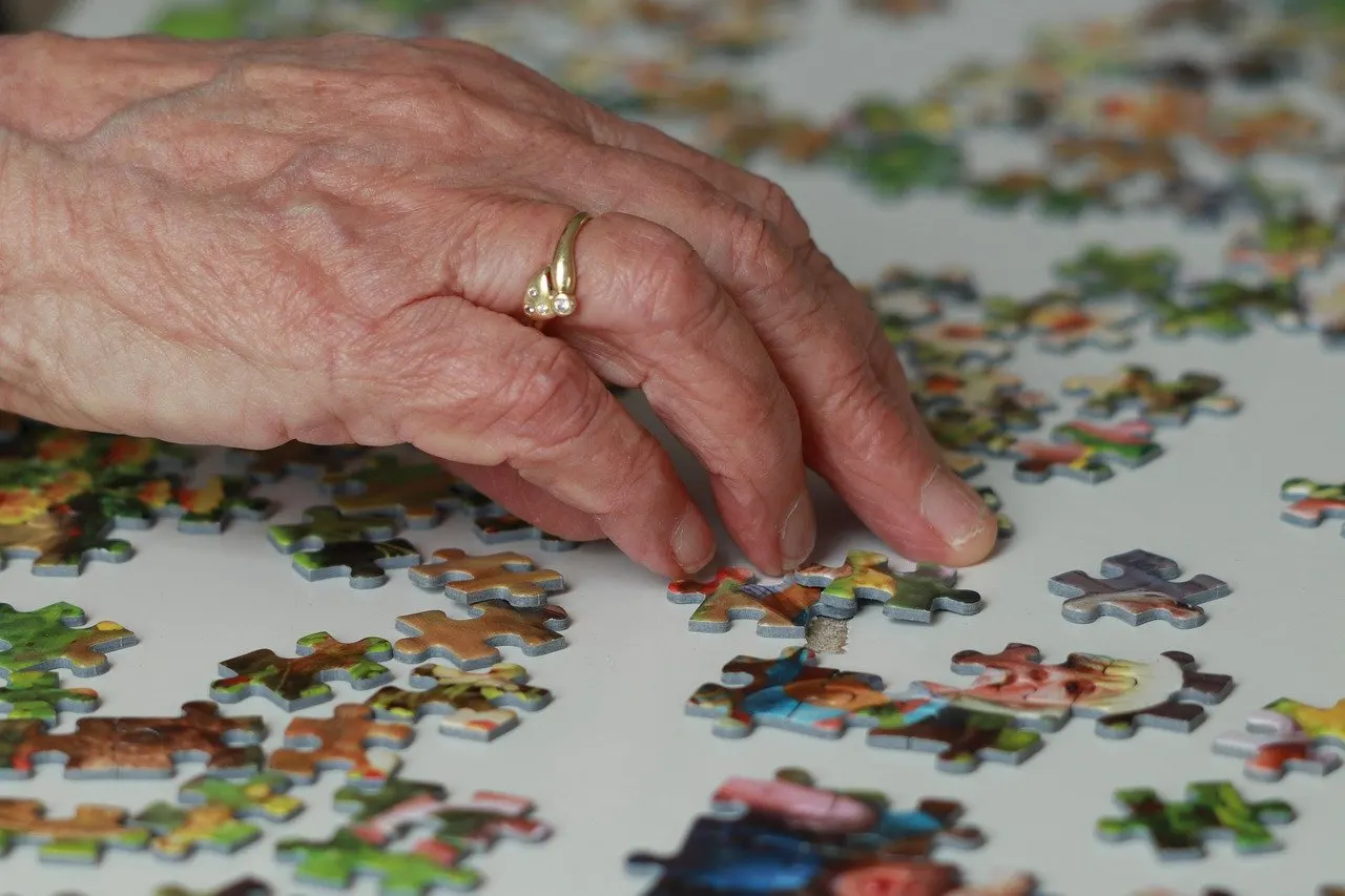 Eine ältere Hand mit goldenem Ring legt ein Puzzleteil auf einem hellen Tisch, auf dem viele bunte Puzzleteile verstreut liegen